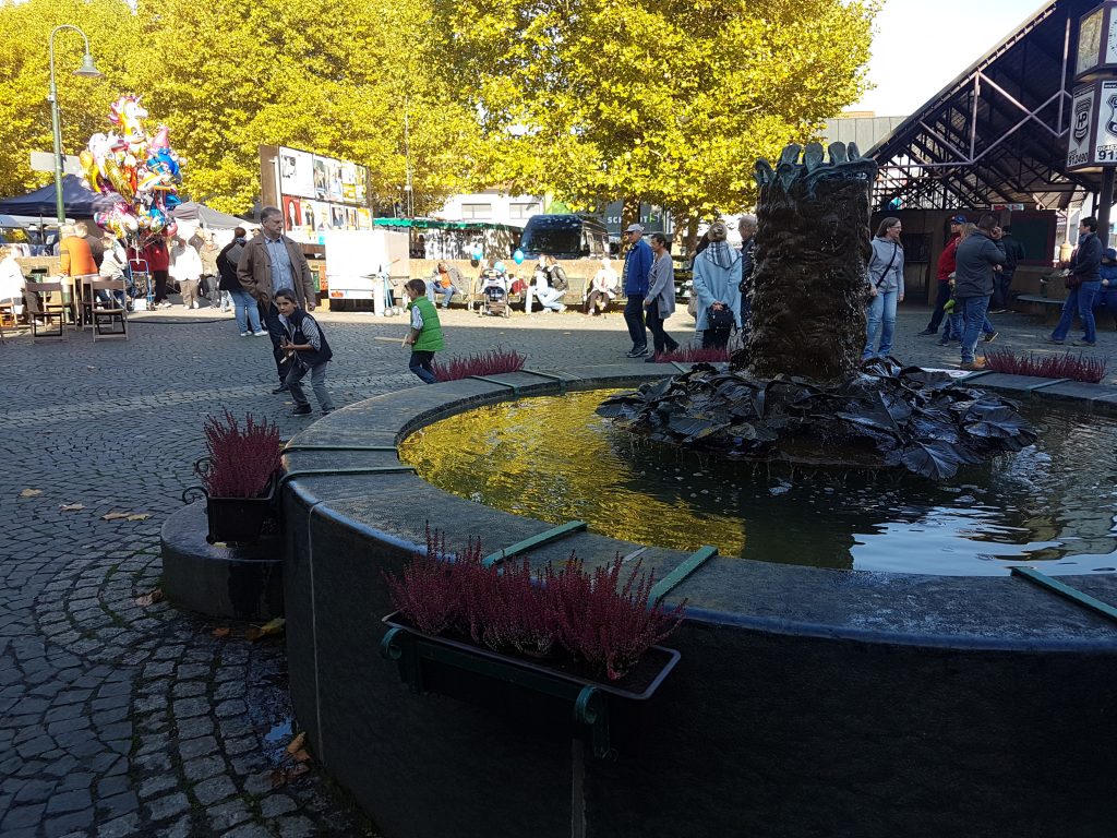Marktplatz mit Brunnen und Leute drumherum.