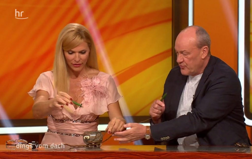 Sonya Kraus und Bodo Bach sitzen am Pult im Studio und rätseln über ein "Dings" von Sibylle Bamberger und untersuchen es gründlich.