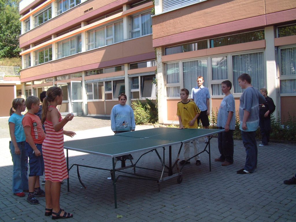 Kinder vor der Schule stehen um die Tischtennisplatte. Sie spielen "Rundlauf".