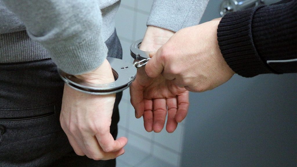 Mann mit Handschellen - es sind nur die Hände zu sehen die auf dem Rücken befindlich sind. Ein Polizeibeamter (Hände) befestigt die Handschellen.