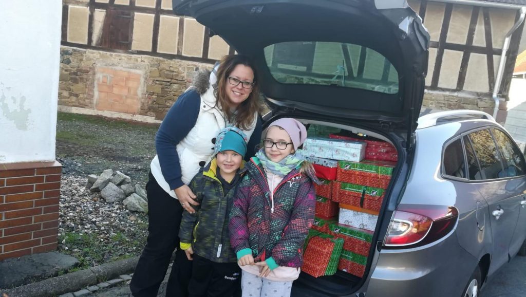 Frau mit 2 Kindern vor einem offenen Kofferraum mit Weihnachtspäckchen