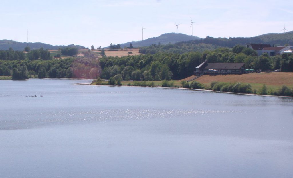 Der Perfstausee. Im Hintergrund die Berghöhen mit Windrädern. Rechterhand das Restaurant Seeblick.