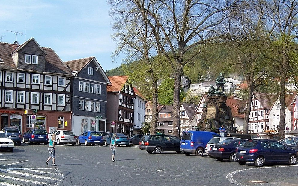 Marktplatz in Biedenkopf, umrahmt von Fachwerkhäusern. Mittendrauf das Kriegerdenkmal mit Brunnen. Parkende Autos überall.