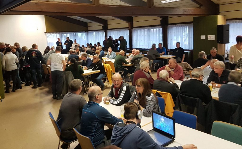 Im Dorfgemeinschaftshaus sitzen die Leute zusammen vor Tischen und genießen das Miteinander bei Getränken und Gesprächen.