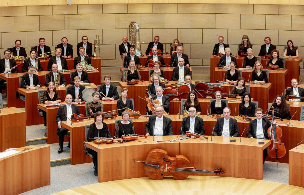 Die Musikerinnen und Musiker der Philharmonie mit ihren Instrumenten. Sitzend, aber nicht spielend.