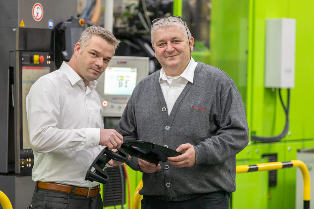 Timothy Schneider (li.), Projektleiter bei Roth Plastic Technology und Dieter Burmeister, Geschäftsführer bei SB Meissner, überprüfen ein Gussteil. Im Hintergrund eine grüne Spritzgussanlage.