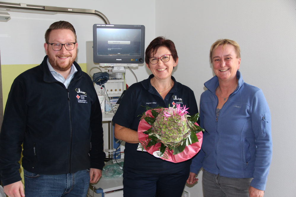 Drei Mitarbeiter des DRK in einem Krankenzimmer mit Überwachungsmonitor, in der Mitte mit Blumenstrauß die neue Atemtherapeutin.