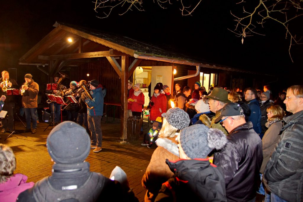 Die beleuchtete Schutzhütte. Davor viele winterlich gekleidete Menschen die dem Posaunenchor zuhören.