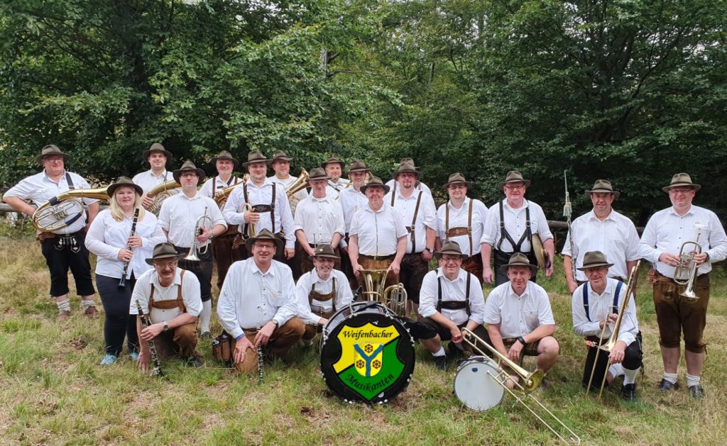 Die Weifenbacher Musikanten - alle im weißen Hemd, mit Lederhosen und Hüten und mit Instrument hocken und stehen als Gruppe auf der Wiese.