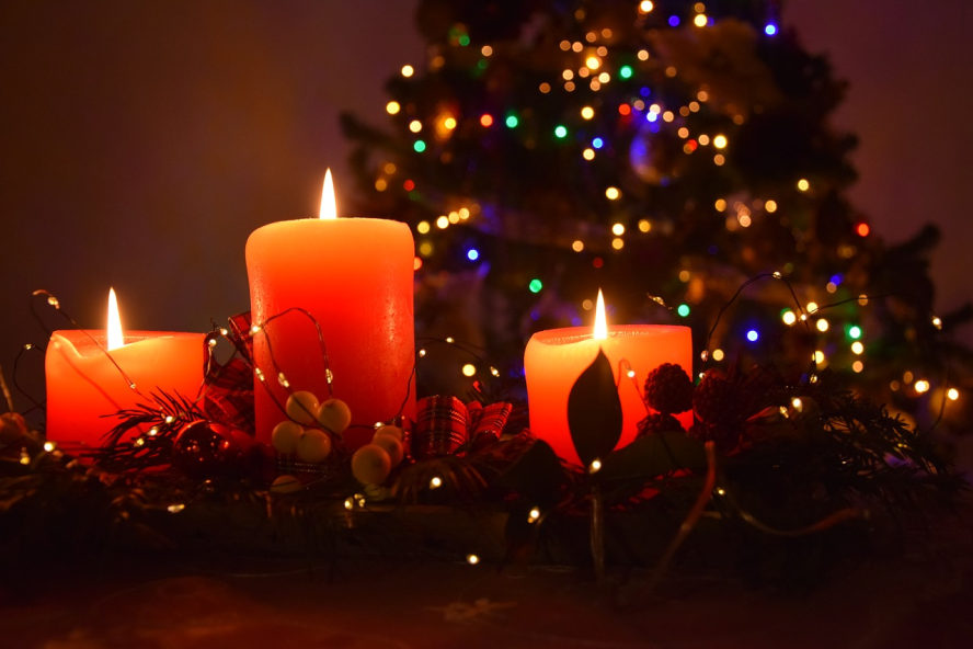 Drei brennende, rote Kerzen. Im Hintergrund ein Weihnachtsbaum zu erahnen. Bunte Lichter daran.