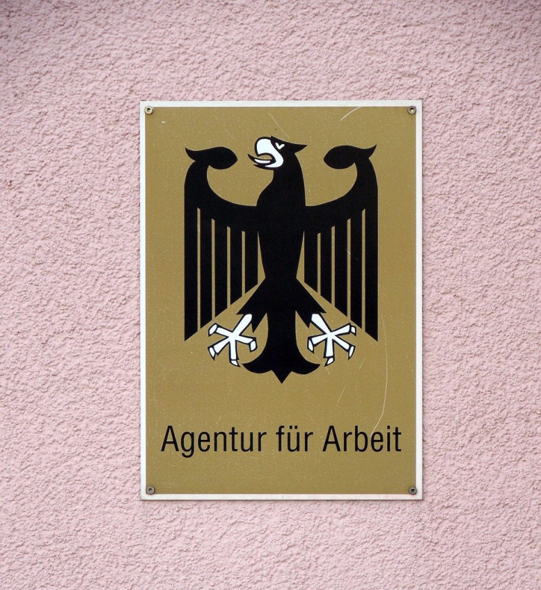 Schild mit Logo (Bundesadler) der Agentur für Arbeit auf rosa Putz-Untergrund.