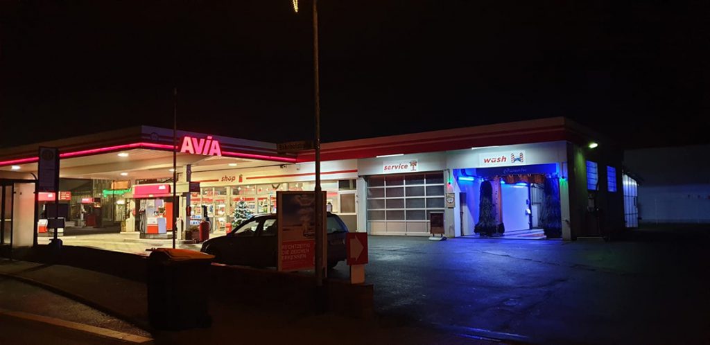 Tankstelle mit angeschlossener Waschanlage und Werkstattbereich, nachts im Dunklen, Tankstelle selbst hell erleuchtet mit Leuchtschrift "AVIA".