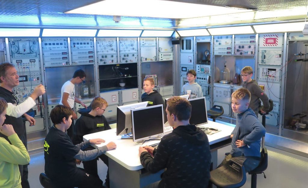 Die Schüler des GSW sitzen an einem Tisch im Raumfahrtlabor und haben Rechner vor sich. Im Hintergrund technische Anlagen undefinierbarer Art.