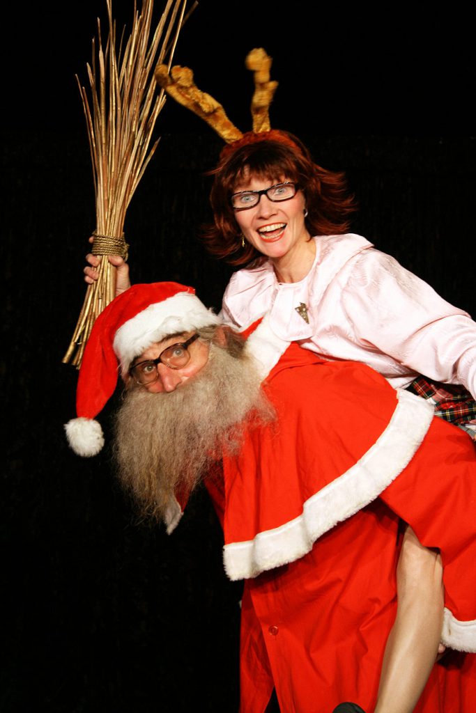 Ein Weihnachtsmann und eine als Rentier verkleidete Frau mit einer Rute in der Hand lachen in die Kamera.