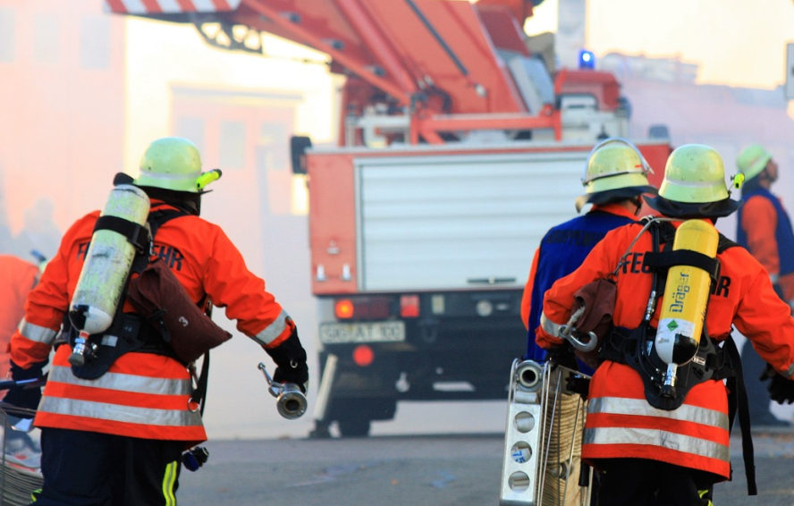 4 Feuerwehrmänner, teils mit Atemschutzgeräten, alle in voller Montur, davor ein Feuerwehrauto, rennen Richtung Brandherd, bereit zu löschen.
