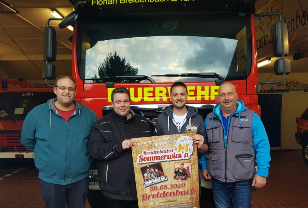 4 Männer stehen vor einem Feuerwehrfahrzeug und halten das Plakat für das Sommerfest.