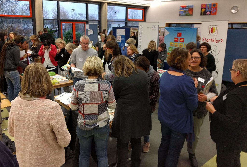 Viele Lehrerinnen und Lehrer in einem Klassenraum diskutieren oder schauen in Prospekte hinein.
