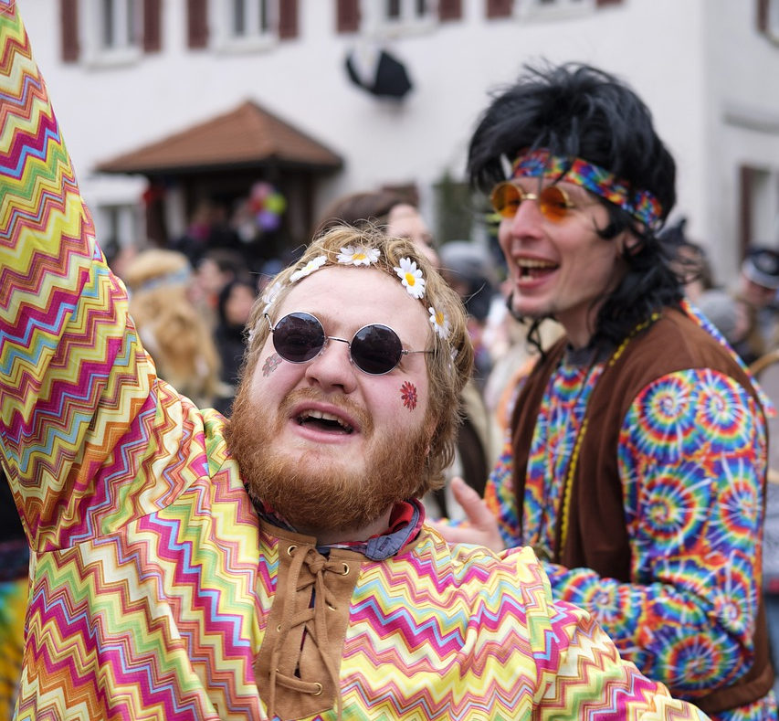 2 wie Hippies gekleidete junge Männer mit Blumenstirnband und bunten Oberteilen, tanzen ausgelassen.