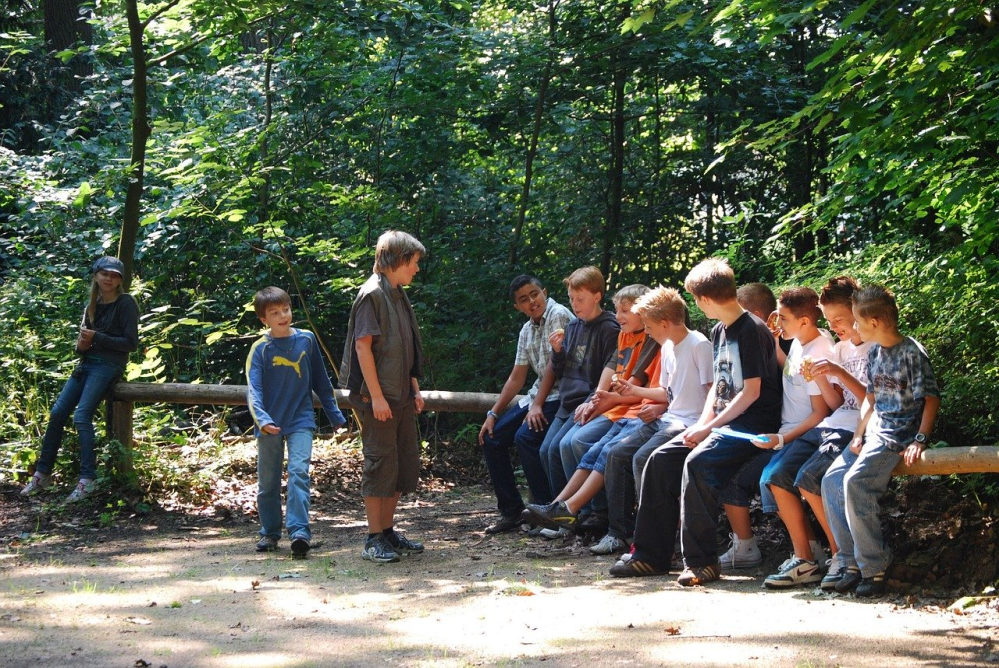 Neun Jungs im Alter zwischen 11 und 14 Jahren sitzen auf einem querliegenden Baumstamm. Zwei stehen davor, ein Mädchen lehnt an einem Baum.