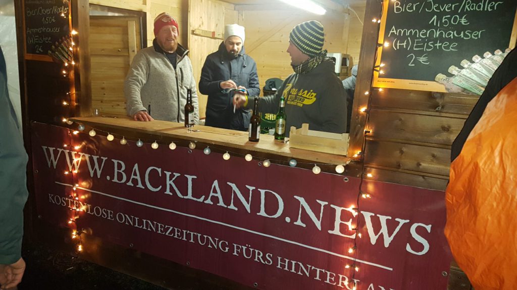 Eine Holzbude, dahinter drei bemützte Fußballvereinsmitglieder. Die Backland.News-Werbung ziert die Front der Bude.