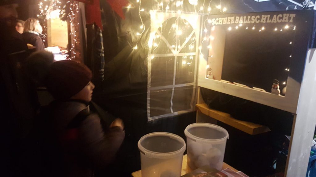 Ein Weihnachtsmarktstand mit einer Wurfbude mit dem Titel "Schneeballschlacht". Der Stand ist von Lichterketten umrahmt.
