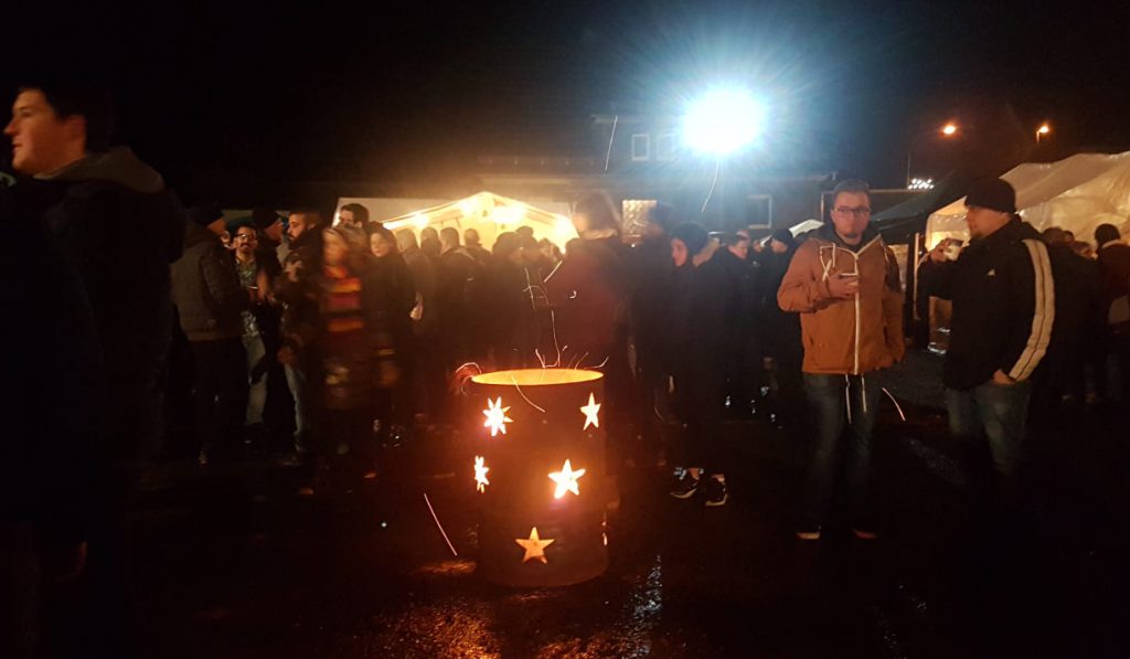 Ein Holz-Feuertopf im Vordergrund und dahinter viele Weihnachtsmarktbesucher mit Glühweintassen oder Snacks in der Hand.