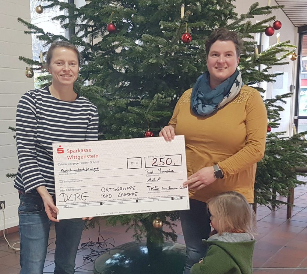 Zwei Frauen vor geschmücktem Tannenbaum mit überdimensioniertem Scheck in der Hand, der die Summe von 250 Euro ausweist.