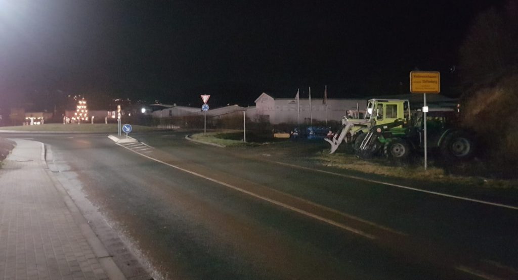 Kreisel bei Niedereisenhausen, rechts das Ortseingangsschild, davor geparkte Traktoren eines benachbarten Fachgeschäfts. Es ist dunkel.