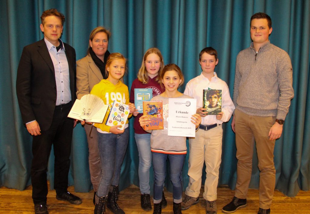 Ein Schüler und drei Schülerinnen mit Buchpreisen in der Hand. Daneben Rektorin und zwei Lehrer. Alle stehen auf der Bühne vor petrolfarbenem Vorhang.