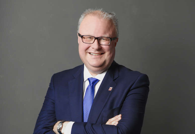 Minister im dunklen Anzug mit verschränkten Armen lächelnd vor neutralem Hintergrund.