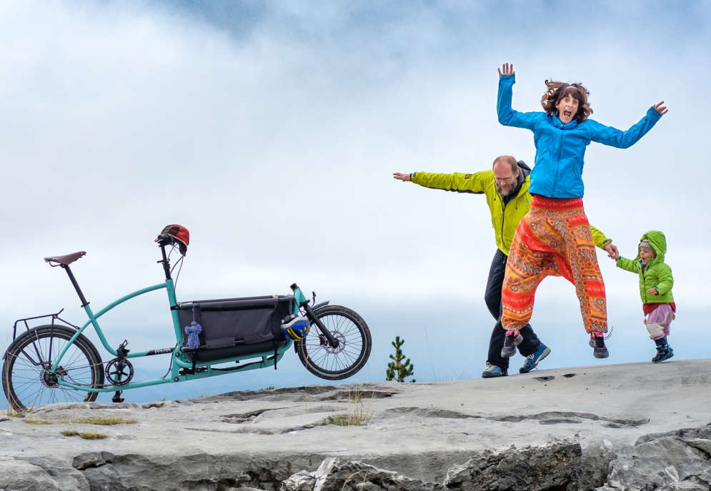 Ein Fels vor dem Meer, ein Lastenrad, eine Familie mit Vater, Mutter, Kind die fröhlich springen.