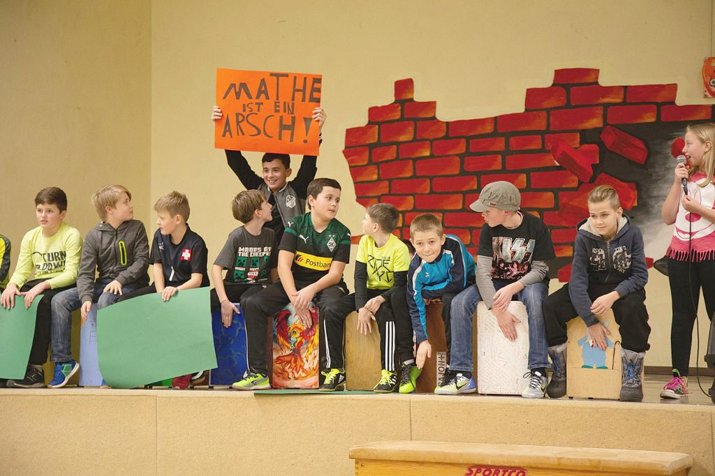 Schülerinnen und Schüler sitzen auf Cajons, singen und halten ein rotes Plakat hoch. Aufschrift: "Mathe ist ein Arsch".