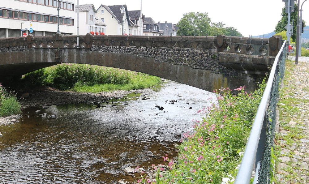 Baufällige Steinbrücke über die Dill die wenig Wasser führt. Rechts ein gepflasterter Weg mit Geländer. Links Gebäude.