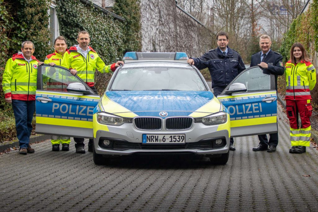 Polizeiauto mit offenen Türen. Daneben zwei Polizisten und vier Notfall-Sanitäter.