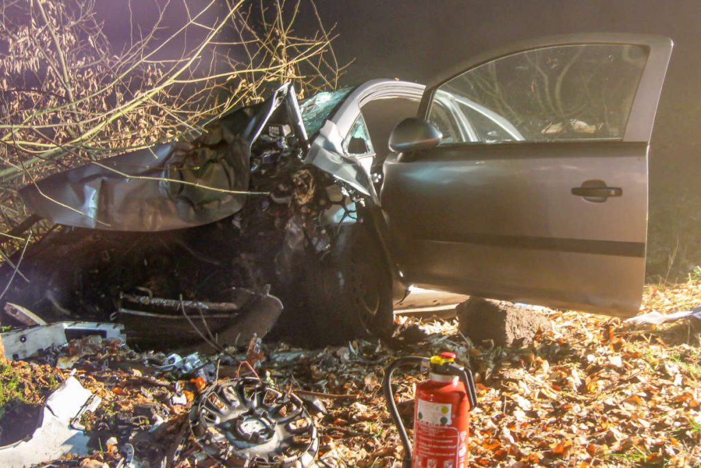 Stark unfallbeschädigter, silberfarbener PKA am Straßenrand. Davor Autoteile und ein Feuerlöscher.