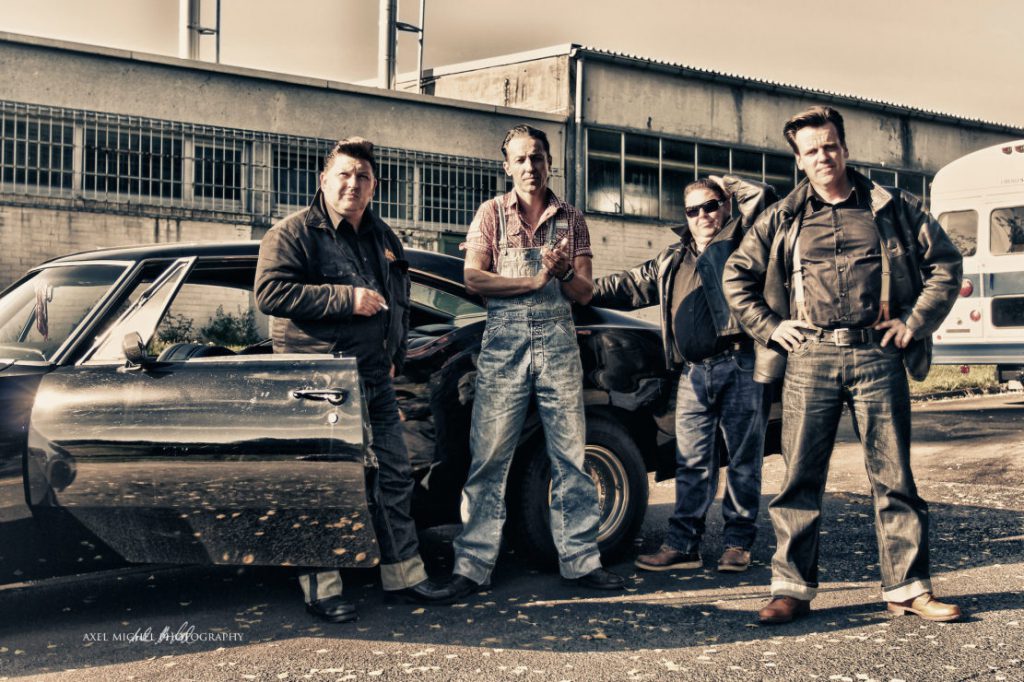 Das Foto mutet
50er Jahre-mäßig an. Vier Jungs stehen in Jeans und Hemden, teils Lederjacken an ein altes Auto gelehnt vor einem alten Fabrikgebäude.
