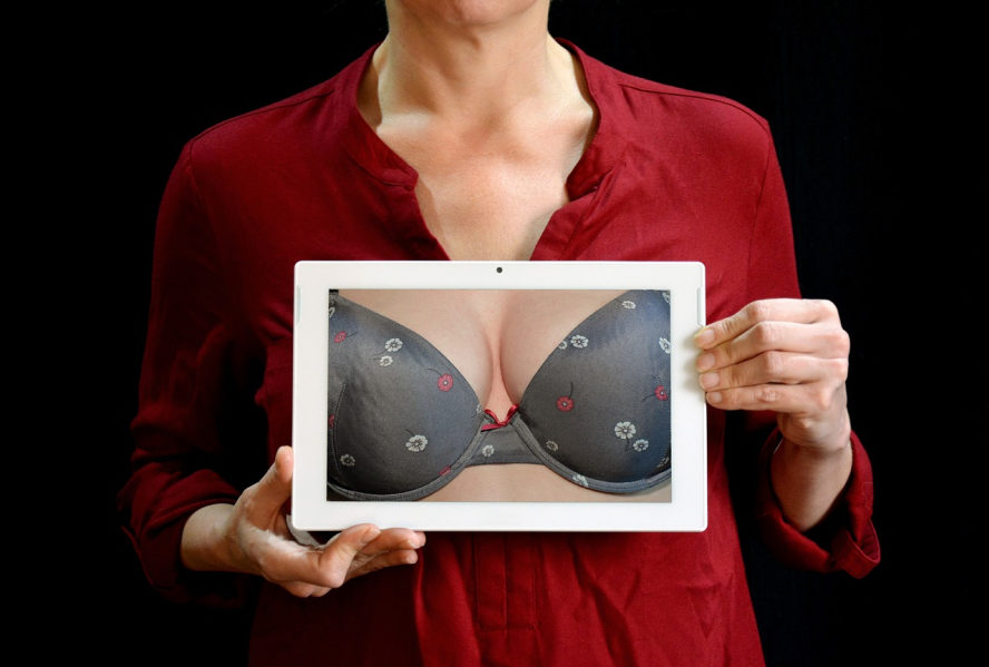 Eine mit einer roten Bluse bekleidete Dame (nur Oberkörper, ohne Kopf) hält ein Tablet vor sich das ein Dekolleté zeigt.