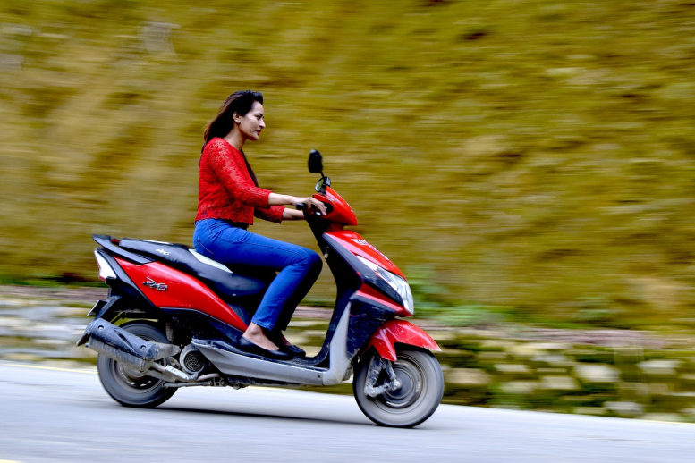 Junge Frau fährt Motorroller. In Jeans und roter Lederjacke, aber ohne Helm. Der Hintergrund ist verschwommen.