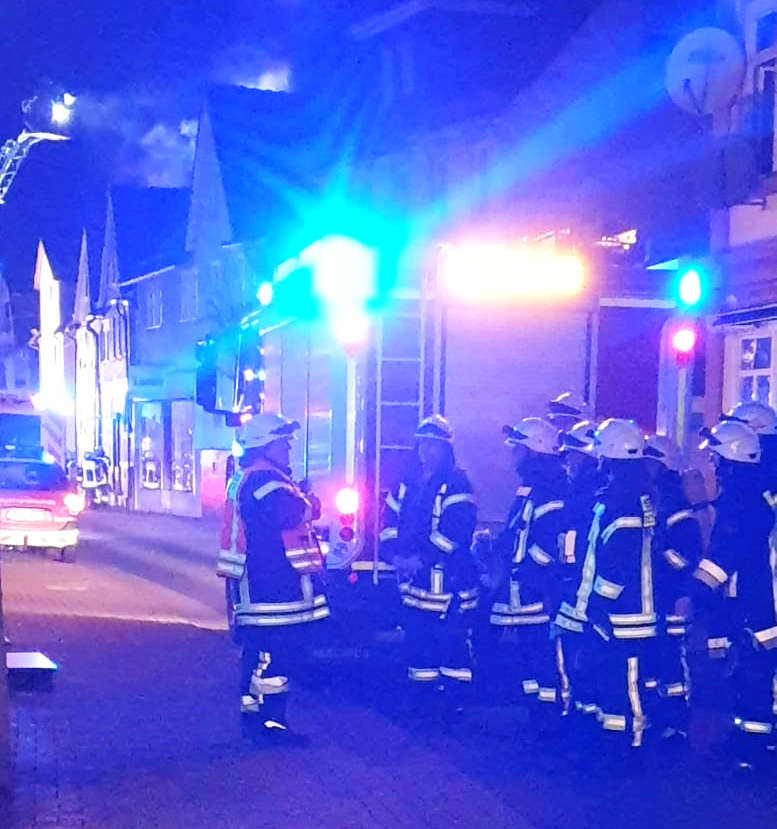 Feuerwehrauto, Einsatzkräfte in Montur. Häuser dahinter.