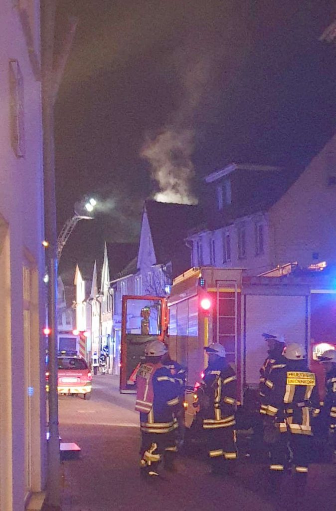 Feuerwehrleute steigen aus dem Einsatzfahrzeug. Hinter einem der Häuser am Straßenrand steigt Rauch auf.
