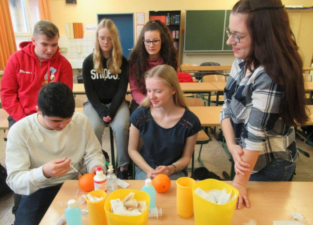 4 Schülerinnen und 2 Schüler sitzen um einen Tisch und ein Junge injiziert eine Flüssigkeit in eine Orange. Die anderen schauen zu.