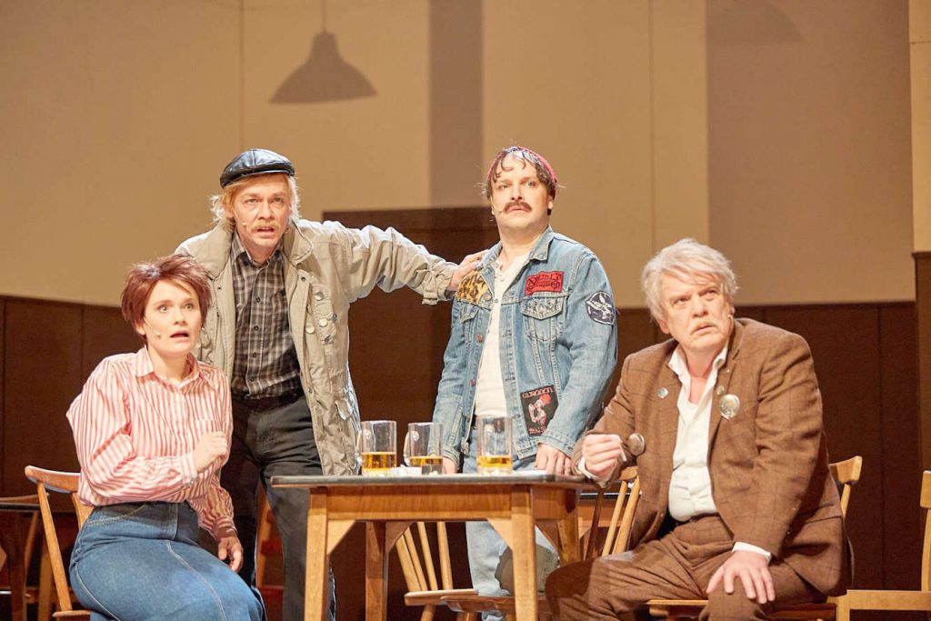 Vier Leute um einen Tisch herum: Schauspieler auf einer Theaterbühne.