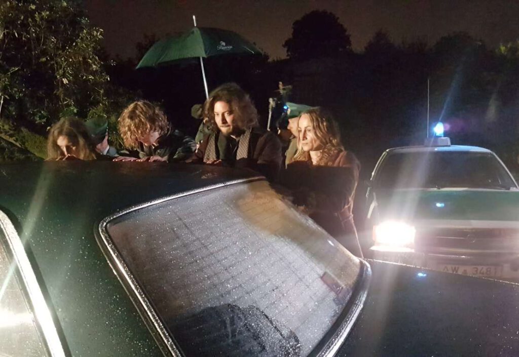 Vier Verdächtige stehen an einem Polizeifahrzeug der 70er Jahre.