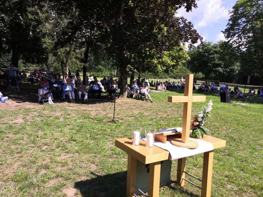 Altar mit Kreuz, Kerzen, Bibel auf einer Wiese. Dahinter auf Bänken und Klappstühlen die Gottesdienstbesucher
