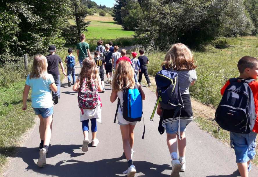 Kindergruppe wandert durch die Wiesen auf geteertem Weg. Alle von hinten zu sehen.