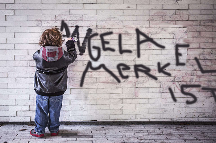 Ein Kind sprüht Graffiti an eine Mauerwand. "Angela Markel ist..."