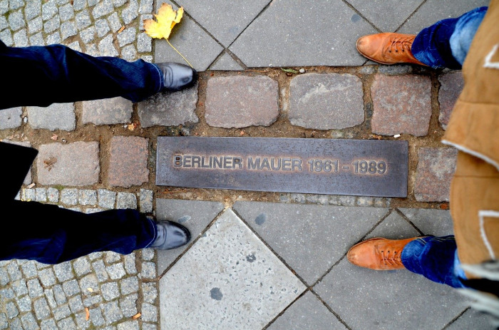 2 Menschen stehen an der Stelle an der früher die Berliner Mauer stand. Eine Gedenktafel ist im Boden eingelassen.