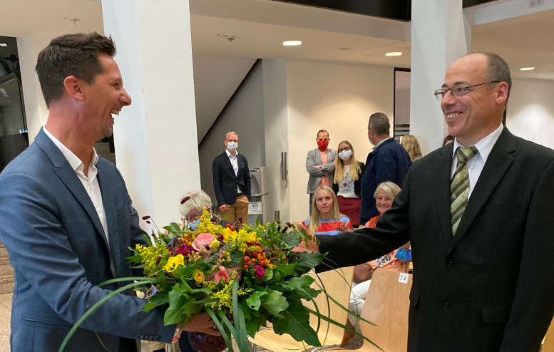 2 Männer lachen miteinander. Einer reicht einen Blumenstrauß