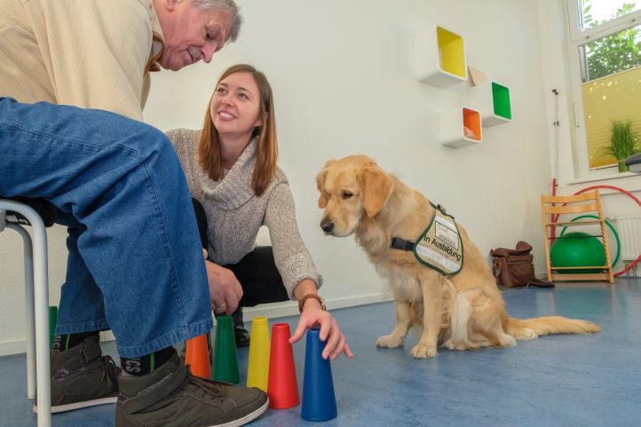 Mensch auf einem Stuhl, davor Ergotherapeutin und Therapiehund.